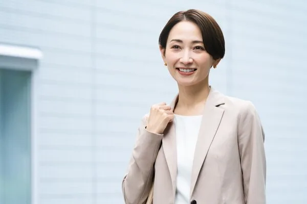 ベージュのビジネスジャケットを着た女性が笑顔で立つ
