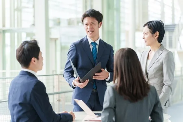 黒いファイルを開いて話す男性と同僚3人