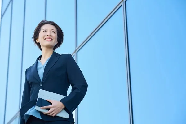 紺スーツとライトブルーシャツの女性がタブレットを持ち、ビル背景で上を見上げている
