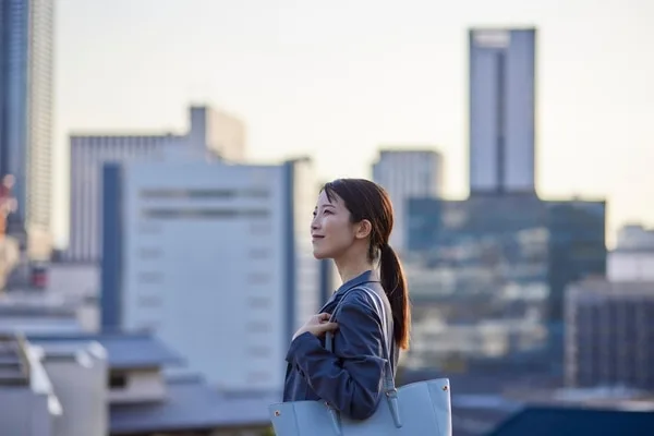 ビジネスバッグを持つ女性、高層ビル群を背景に遠くを見つめる
