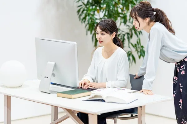 オフィスのデスクでパソコン画面を見ながら営業資料について打ち合わせする二人の女性