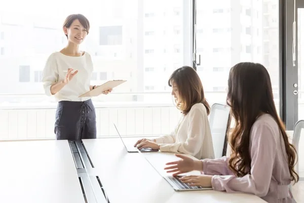 オフィスで営業女性がクリップボードを持ち、2名の女性に説明している