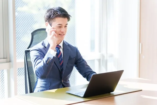 ビジネススーツの男性がデスクでノートパソコンを操作しながら電話で通話中
