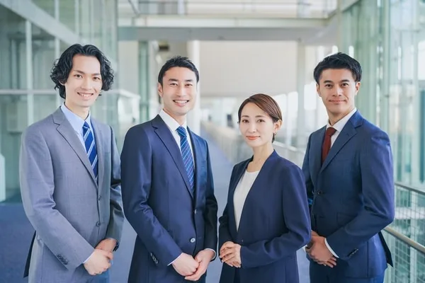 ビジネススーツの営業チーム4人がオフィスの廊下で笑顔で立つ