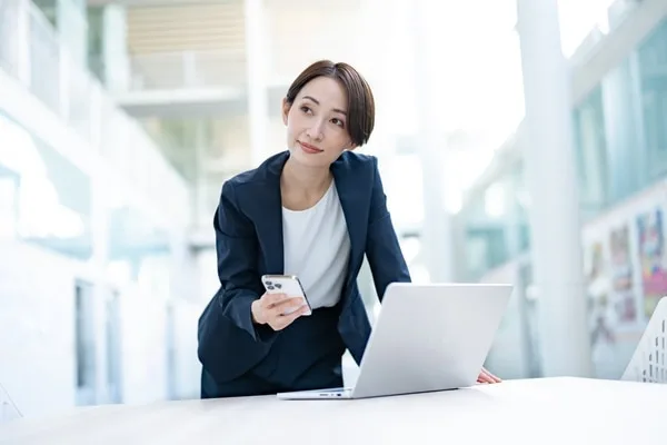 ノートパソコン前でスマートフォンを持つ女性