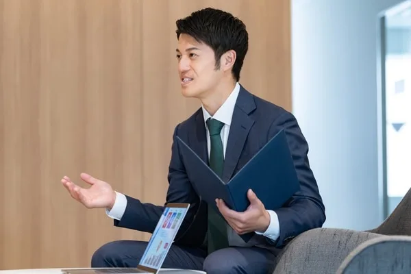 バインダーを手に身振りで説明するスーツ姿の男性