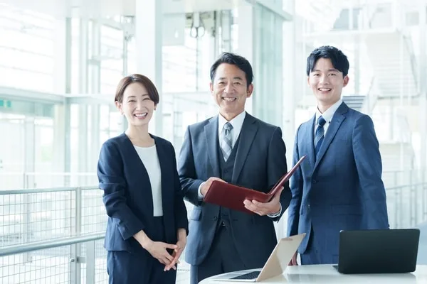 赤いファイルを開き笑顔で並ぶスーツ姿の3人