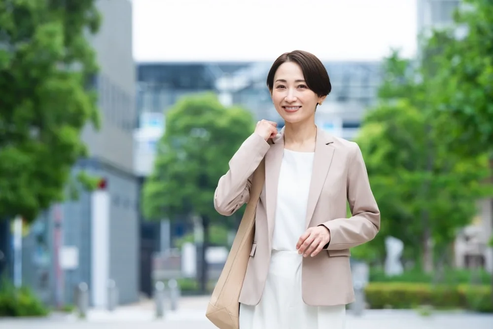 ベージュのジャケットを着た女性が街中で笑顔で立つ