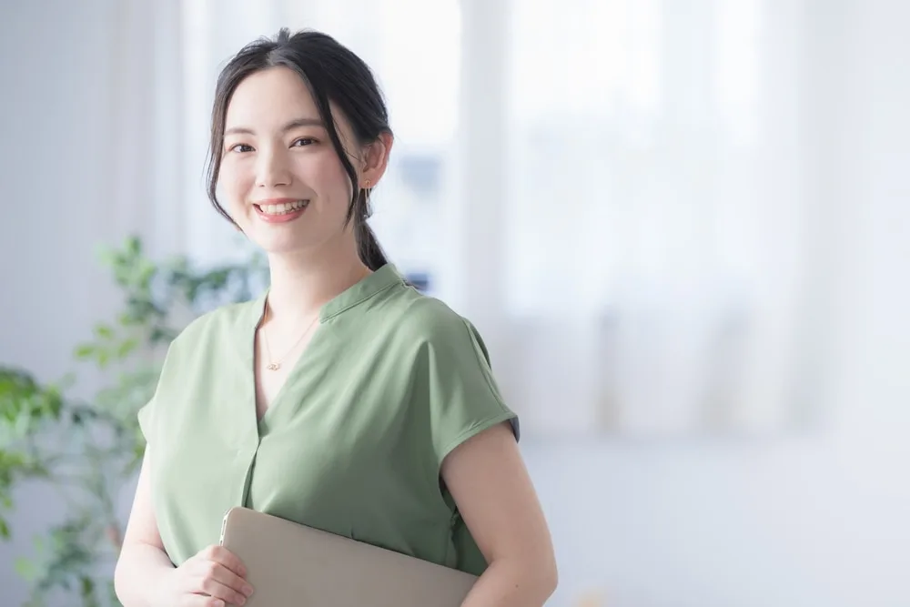 緑色のシャツを着た女性がタブレットを持ってオフィスで笑顔で立つ