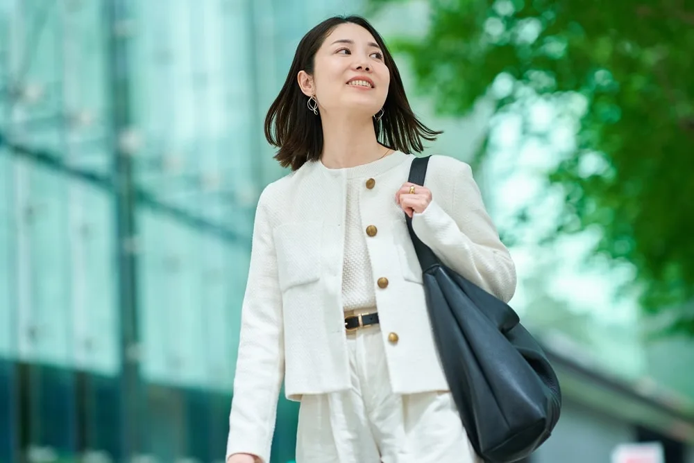 白いジャケットとパンツを着た女性が黒いバッグを肩にかけ上を向いて笑顔
