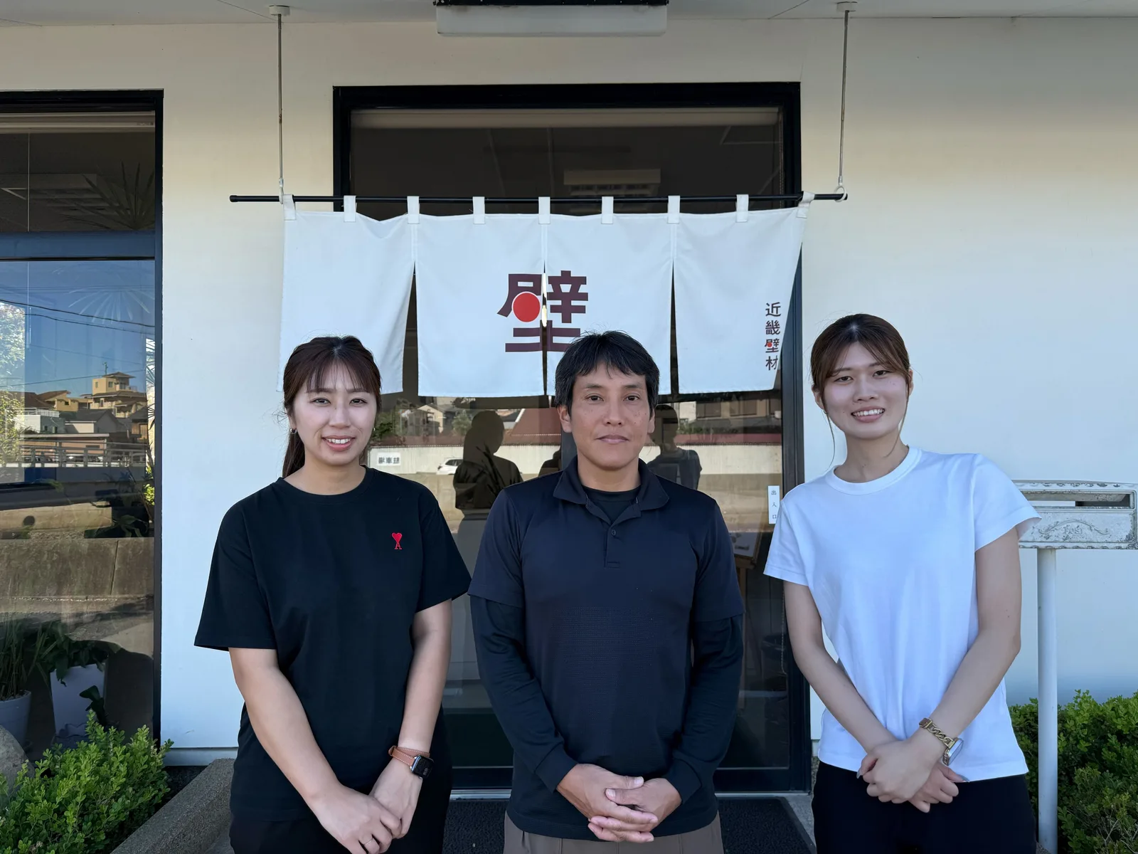 近畿壁材工業の店舗前で笑顔で並ぶ三人のスタッフ