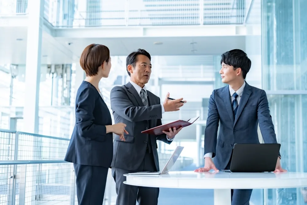 ノートパソコンを置いた机で立ち話するスーツ姿の3人