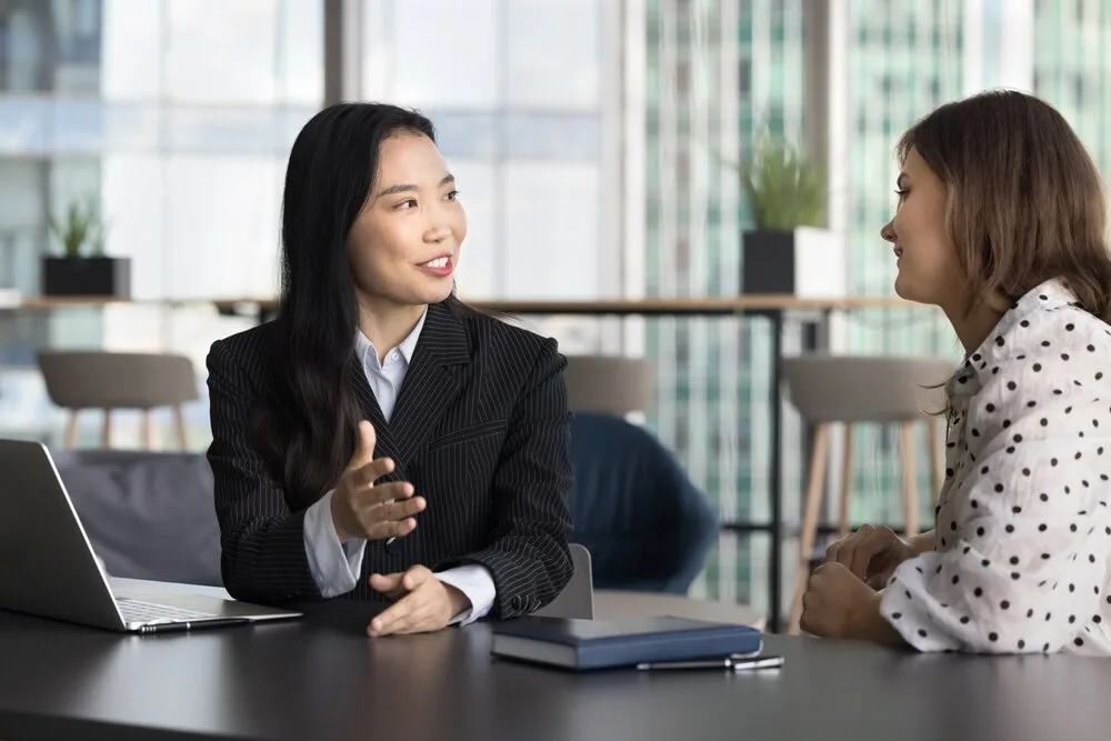 オフィスの机でノートパソコンを前に会話する二人の女性ビジネスパーソン