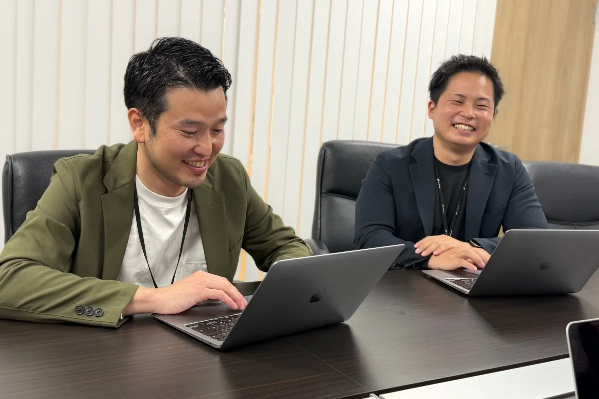 複数人が会議テーブルでノートパソコンを操作しながら会話している場面