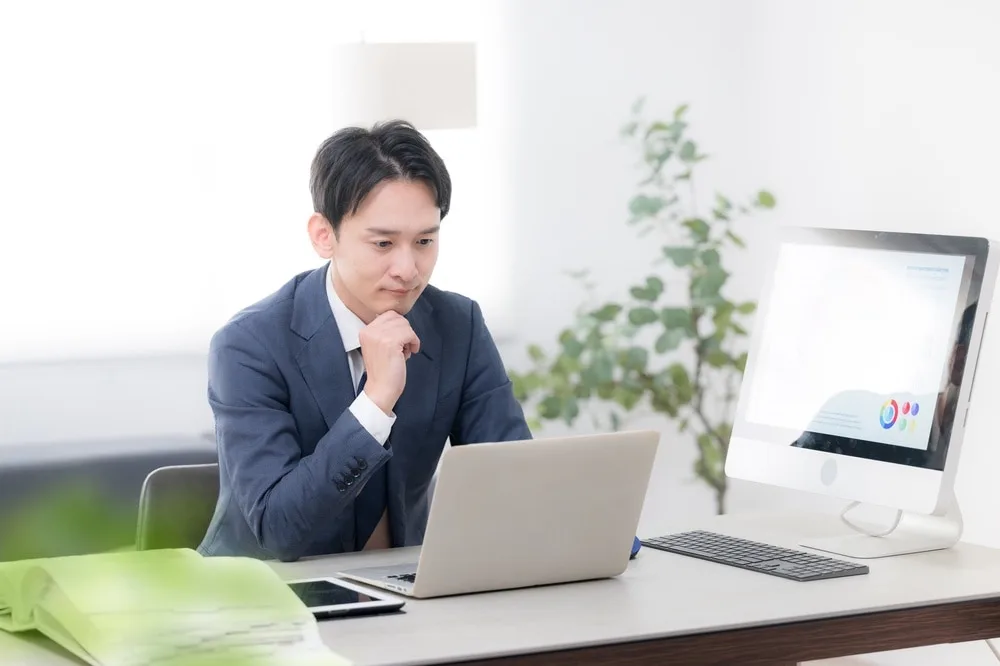 ノートパソコンとデスクトップディスプレイの前で資料を確認する男性営業担当者