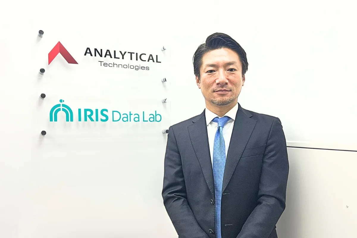 IRISデータラボ株式会社のロゴ看板の横に立つ、スーツ姿の取締役・中塚様の全身カット