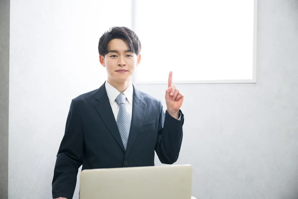 ビジネススーツの男性がノートパソコン前で人差し指を立て指摘するジェスチャー