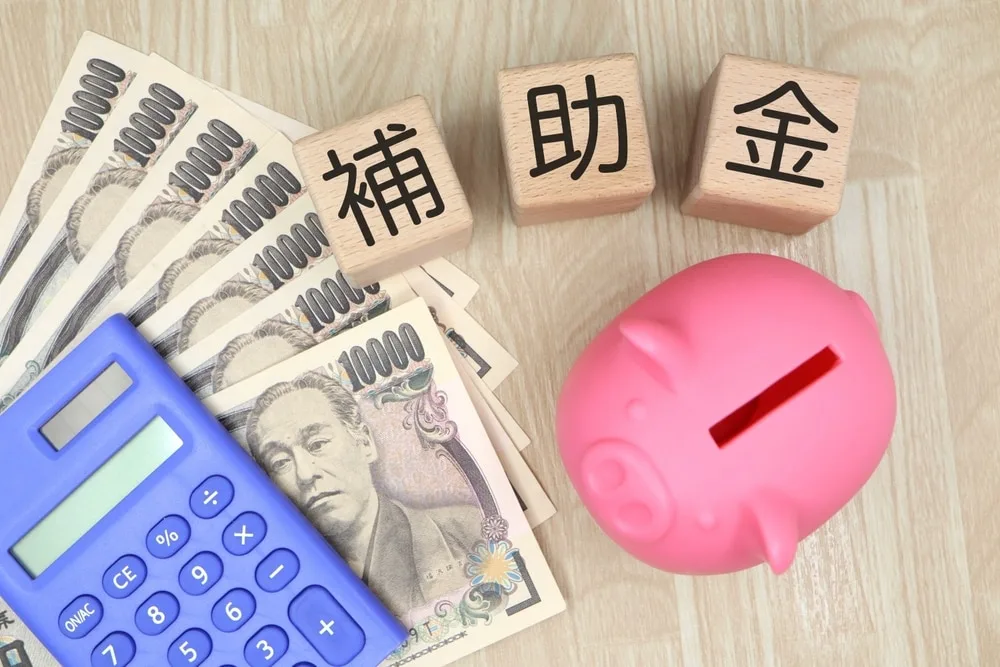 補助金の木製ブロックと一万円札、電卓、豚の貯金箱
