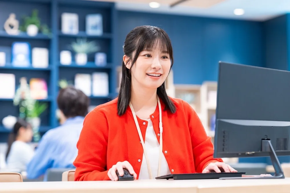 赤いジャケットの女性がデスクでパソコンを操作している
