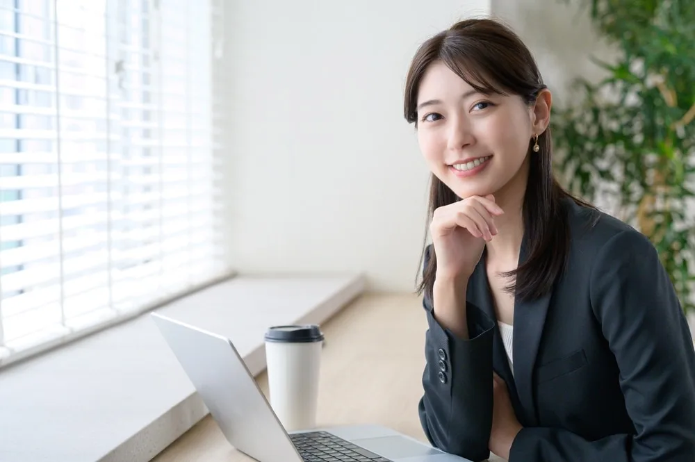 オフィスのデスクでノートパソコンを前に笑顔でポーズする女性