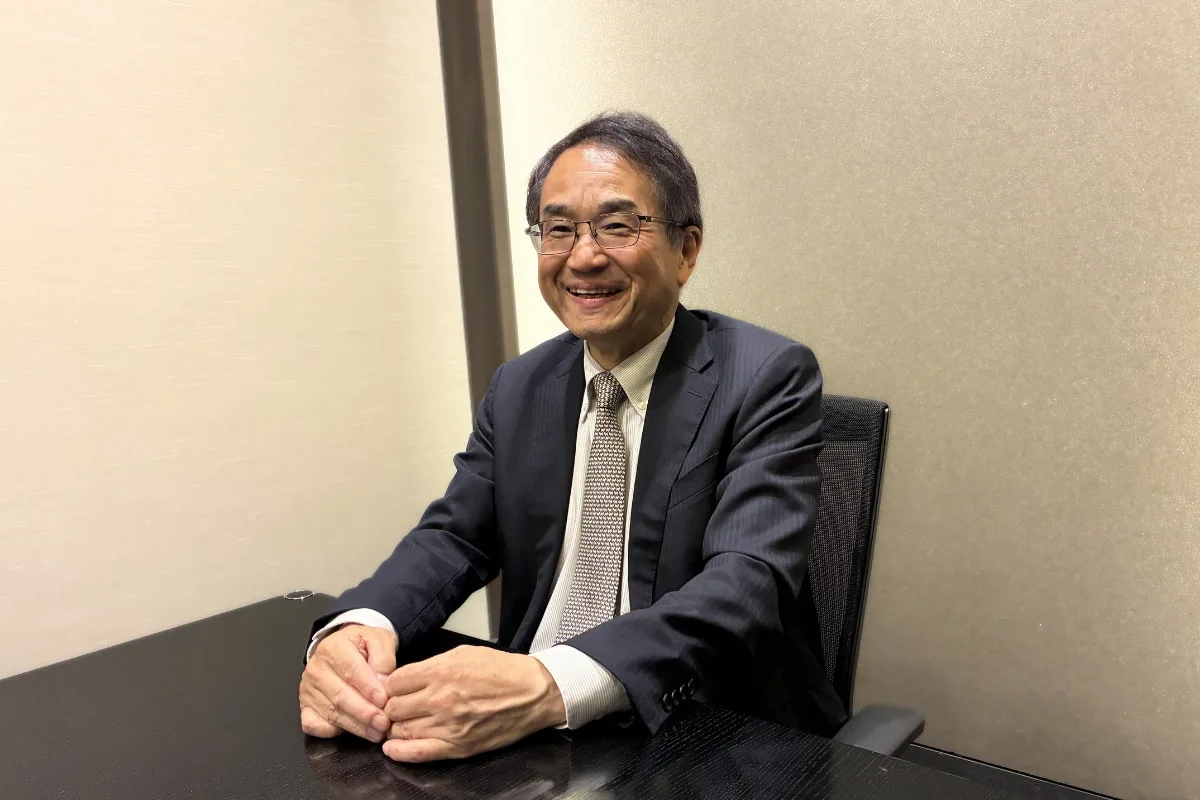 紺色スーツを着た男性が机に向かって笑顔で座っている